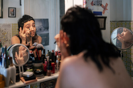 Back view alternative topless female applying eyeliner while sitting at vanity table and reflecting in mirror