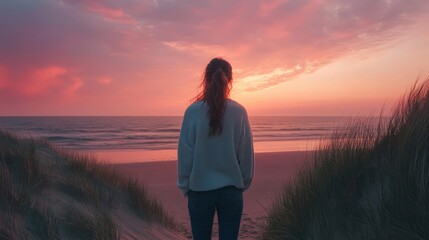 A serene moment at sunset, a woman gazes at the calm ocean waves and colorful sky, surrounded by gentle grass on the beach.