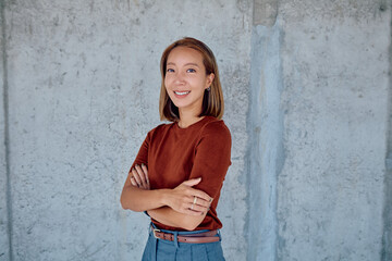 Portrait of a smiling young Asian businesswoman standing with arms crossed against a concrete wall,...