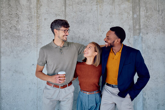 Three cheerful multi-ethnic colleagues standing together with arms around each other's shoulders, smiling and enjoying a coffee break