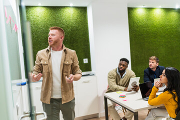 Young manager is leading a business meeting with his team, presenting a project in a modern...