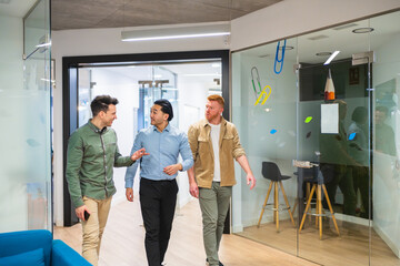 Three young male professionals walking and talking in a modern coworking office, discussing business matters in a collaborative environment. One is chinese