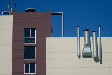 Silver ventilation pipes and ducts affixed to a pale siding facade flank a dark‑tinted window,...