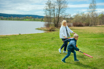 A young grandmother and her grandson playing badminton on a green meadow near a mountain lake....