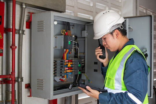 Engineer conducts fire safety inspection near electrical distribution board while communicating on walkie talkie and holding tablet in safety helmet and reflective vest