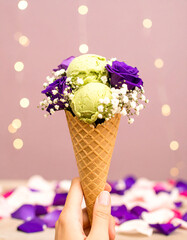 Elegant artistic photo of waffle cone filled with green matcha ice cream, surrounded by violet roses and delicate baby’s breath flowers
