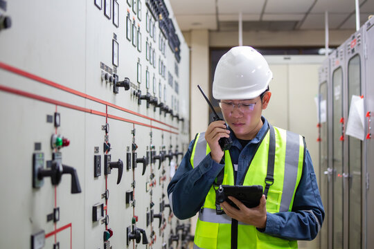 Power station engineer in control room wearing safety helmet and reflective vest using walkie talkie and tablet for monitoring electrical system with focused expression