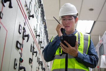 Power station engineer in control room wearing safety helmet and vest using walkie talkie and...