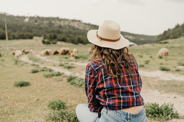 A woman in a plaid shirt and hat is sitting in a field with a herd of sheep
