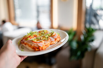 Hand holding a white plate with two fresh slices of pizza topped with ham, tomatoes, and greens,...
