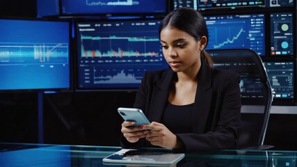 Young female financial analyst using smartphone and multiple computer screens showing charts, graphs, and data in a dark office at night, working late and analyzing market trends - Powered by Adobe