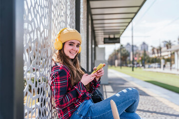 Smiling female student using smartphone while listening music at public transport stop