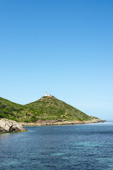 A serene coastal view of a green hill in Turkiye, with a lighthouse at the top, under a clear blue...