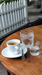 A cup of hot espresso served with a glass of melted sugar, and a large glass of ice water on a wooden table looks luxurious. Natural blur background, in outdoor cafe.