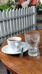 A cup of hot espresso served with a glass of melted sugar, and a large glass of ice water on a wooden table looks luxurious. Natural blur background, in outdoor cafe.