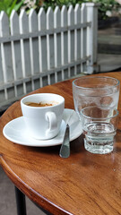 A cup of hot espresso served with a glass of melted sugar, and a large glass of ice water on a wooden table looks luxurious. Natural blur background, in outdoor cafe.