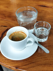 A cup of hot espresso served with a glass of melted sugar, and a large glass of ice water on a wooden table looks luxurious. Natural blur background, in outdoor cafe.