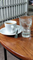 A cup of hot espresso served with a glass of melted sugar, and a large glass of ice water on a wooden table looks luxurious. Natural blur background, in outdoor cafe.