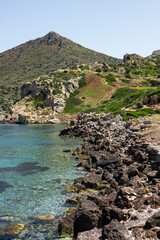 A rocky coastline in Turkiye with clear turquoise water and a green hillside. A mountain rises in...