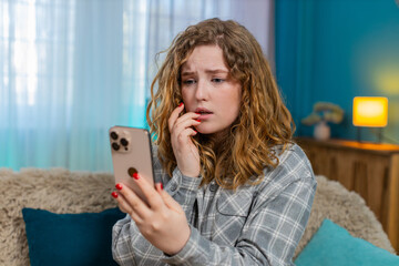 Young woman at home anxiously waiting for important message biting nail while scrolling smartphone....