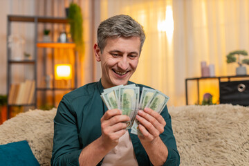 Young man at home counts cash dollars with wide smile, clearly satisfied and pleased with gain....