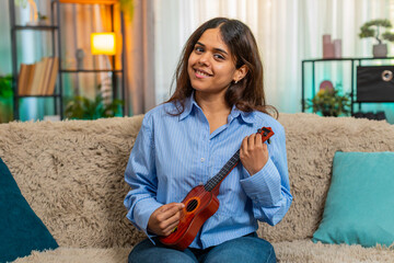 Indian woman plays toy ukulele on home sofa, laughing with joy and pretending to perform like...