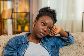 African American young woman looks deeply stressed while sitting alone on couch, reflecting on an...