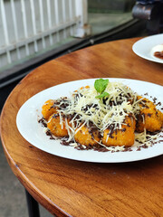 A pile of crispy fried bananas covered with grated cheese and chocolate sprinkles, decorated with fresh mint leaves, served on an oval white plate on a wooden table, in an outdoor cafe.
