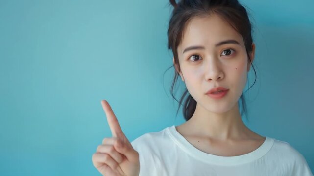 Young Woman Pointing Up Against Blue Background - A young Asian woman with dark hair tied back in a low bun is shown from the chest up against a solid light blue background.