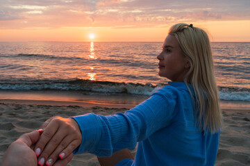 A romantic evening on the seashore. A beautiful blonde woman holds her beloved's hand, admiring the...