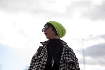 A fashionable woman sporting a vibrant green hat and stylish sunglasses is standing confidently in...