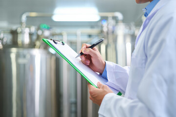 Scientist in lab coat writing on clipboard in laboratory with large tanks in the background