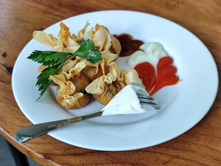 Crispy fried dumplings served with celery leaves, as well as chili, tomato and mayonnaise sauce arranged artistically on a white plate on a wooden table in an outdoor cafe. Natural blur background.
