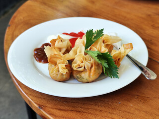 Crispy fried dumplings served with celery leaves, as well as chili, tomato and mayonnaise sauce arranged artistically on a white plate on a wooden table in an outdoor cafe. Natural blur background.