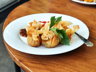 Crispy fried dumplings served with celery leaves, as well as chili, tomato and mayonnaise sauce arranged artistically on a white plate on a wooden table in an outdoor cafe. Natural blur background.