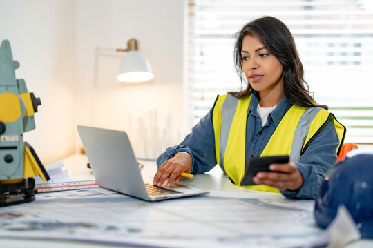 Female construction project manager professional working indoors using laptop wearing  bright safety jacket in modern workspace