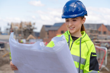 A young female construction worker in a reflective jacket and hard hat is studying blueprints at a...