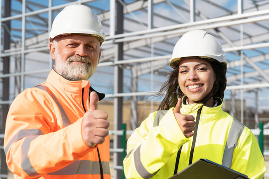 Male and female construction workers giving thumbs-up as they discuss plans on a site while wearing safety gear and reviewing documents