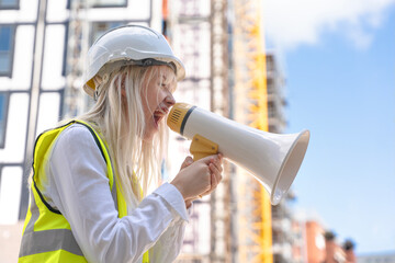 Portrait of young woman construction worker in vivid safety gear uses a megaphone to communicate...