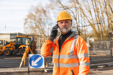 Construction works site manager  in orange safety vest and hard hat speaks on the phone at road...