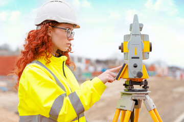Young female surveyor operates a total station at a construction site while wearing safety hi-viz...