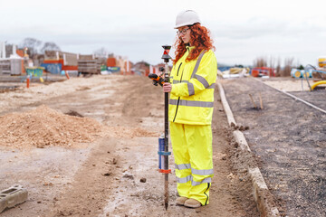 Portrait of confident female land surveyor with long hair in bright safety gear using surveying...