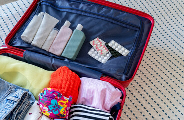 Open suitcase overflowing with clothes on bed in bedroom for a trip. Take with you the necessary pills and medicines, cosmetic accessories, beauty products and clothing. Getting ready for a vacation.