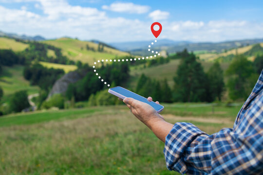 A close-up of a hiker's hands holding a smartphone, tracking a digital route marked by a dotted line leading to a red location pin on the horizon. The image captures the integration of modern