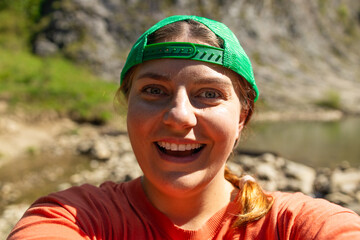 Energetic young woman in a green cap and orange shirt taking a joyful selfie by a mountain stream....