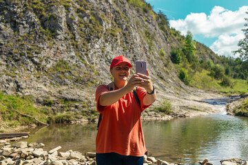 Smiling young woman in casual hiking attire and red cap sitting on a rock and taking a selfie near a peaceful mountain stream. Surrounded by natural beauty and rocky terrain, she enjoys a break during