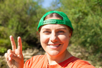 Cheerful young woman in a green cap and orange shirt smiling and showing a peace sign during a hike in nature. Close-up portrait with blurred green background. Ideal for themes of youth, freedom