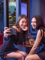 Two young women taking a selfie together at a bar while enjoying drinks. They are smiling happily, dressed stylishly, and sitting at a round table with beer glasses