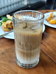 A glass of iced milk coffee with a clear coating, placed on a wooden table, with a slightly blurred background of food dishes, outdoor cafe.