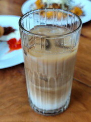 A glass of iced milk coffee with a clear coating, placed on a wooden table, with a slightly blurred background of food dishes, outdoor cafe.